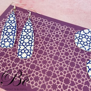 May include: A pair of blue and white geometric patterned dangle earrings with silver hooks. The earrings are on a purple background with a geometric pattern.