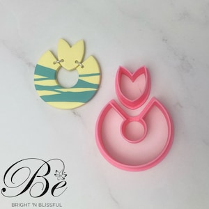 May include: A pink plastic cookie cutter shaped like a flower with a ring in the centre. The cookie cutter is on a white surface next to a yellow and green cookie.