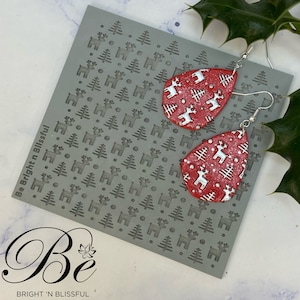 May include: A pair of red teardrop-shaped earrings with a white reindeer and tree pattern. The earrings are on a gray background with a repeating pattern of reindeer and trees.