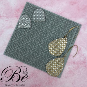 May include: A pair of gold and white teardrop-shaped earrings with a geometric pattern. The earrings are on a gray silicone mat with a repeating pattern. The mat has the text "Be Bright 'n Blissful" printed on it.