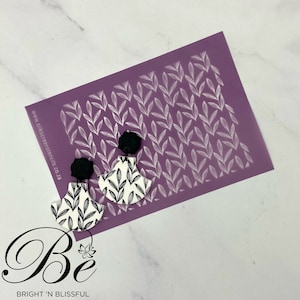 Op de afbeelding: Een paarse stencil sheet met een herhalend patroon van witte bladeren. Twee witte keramische oorbellen met een zwart bladpatroon zijn te zien op een wit oppervlak. De oorbellen zijn gevormd als een halve maan. De tekst "www.silkscreenstencils.co.za" is afgedrukt op de stencil sheet.