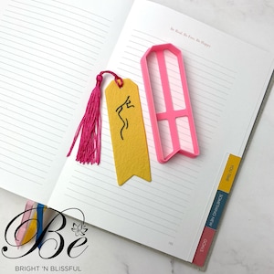 Puede incluir: Un marcapáginas rosa con forma rectangular y un rectángulo más pequeño en el medio. El marcapáginas está en una página blanca con papel rayado. La página tiene el texto "Be Real. Be Free. Be Happy." en la parte superior. La página también tiene el texto "YOU TIME", "SOMETHING NEW" y "GOALS" en el lado derecho. La página es parte de una libreta con el texto "Be BRIGHT 'N BLISSFUL" en la parte inferior.