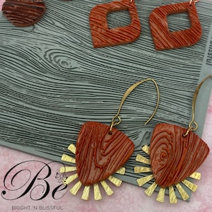 May include: A pair of brown wood-grain textured earrings with gold accents. The earrings are shaped like a triangle with a sunburst design. The earrings are on a gray textured surface.