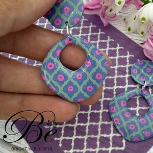 May include: A pair of square-shaped earrings with a geometric pattern in shades of blue, green, and pink. The earrings have a large hole in the centre and are made of polymer clay.