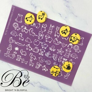 Peut inclure: Cinq emporte-pièces en fondant jaune avec des motifs d'animaux noirs sur un tapis de cuisson en silicone violet. Les emporte-pièces représentent un cheval, un lama, un mouton, un lapin et un chat. Le tapis est imprimé de divers motifs d'animaux. Le texte "www.silkscreenstencils.co.za" est imprimé sur le tapis.
