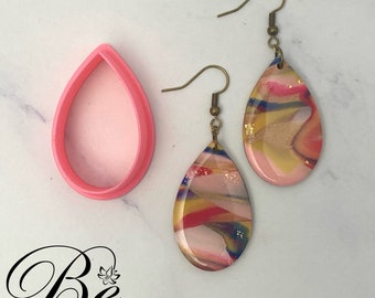 May include: A pair of teardrop-shaped earrings with a colourful abstract design. The earrings are made of resin and have a gold-tone finish. The earrings are shown with a pink plastic mould.