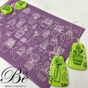 May include: A pair of green and black dangle earrings with a cactus and a flower in a vase design. The earrings are on a purple background with a white pattern of potted plants.