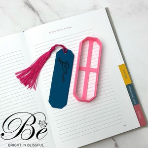 May include: A teal blue leather bookmark with a pink tassel and a black silhouette of a dancer. The bookmark is lying on a page of a journal with the text "Be Real. Be Free. Be Happy." and the words "You Time", "Something New", and "Goals" written on the tabs of the journal.