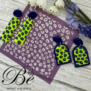 May include: A set of four statement earrings featuring a bright yellow and blue leopard print design. The earrings are made of polymer clay and have a blue glaze. The earrings are displayed on a purple background with a white leopard print pattern.