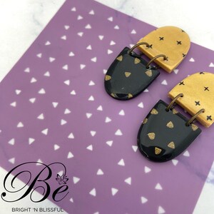 May include: A pair of statement earrings with a black and gold geometric design. The earrings are made of polymer clay and feature a black base with gold accents. The earrings are suspended from a gold-toned metal hook.
