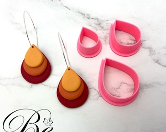 May include: Three pink polymer clay cutters shaped like teardrops, with a pair of earrings made of three layered teardrop shapes in red, orange, and yellow. The earrings are hanging from silver hoops. The text "Be Bright 'n Blissful" is at the bottom of the image.