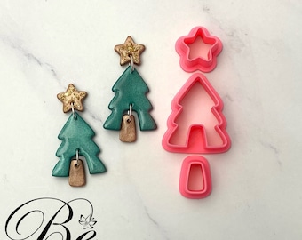 May include: A pair of teal green Christmas tree earrings with gold glitter stars and gold accents. The earrings are shown next to a pink plastic Christmas tree cookie cutter.