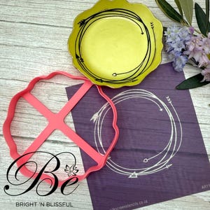 May include: A pink plastic stencil with a circle design and a cross shape in the centre. The stencil is on a purple background with a white circle design with arrows around it. The text "Be Bright 'n Blissful" is at the bottom of the image.