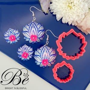 May include: A set of three pink and blue lotus flower earrings and two cookie cutters. The earrings are made of resin and have a silver hook. The cookie cutters are made of pink plastic and are shaped like lotus flowers.