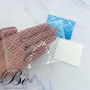 May include: A clear plastic stencil with a geometric pattern of white squares and rectangles. The stencil is being held in a hand over a blue and white patterned surface.