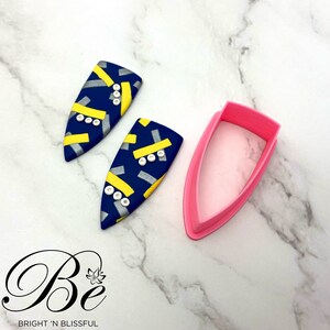 Puede incluir: Dos pendientes de arcilla con estampado geométrico azul, amarillo y plateado con detalles de perlas blancas. También se muestra un cortador de plástico rosa en la imagen.