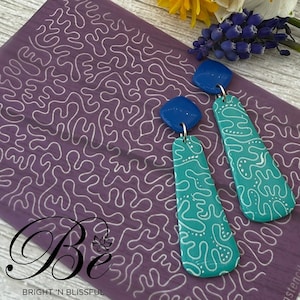 May include: A pair of dangle earrings with a teal blue and white patterned design. The top of each earring is a small, square, royal blue piece.