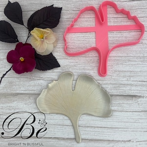 May include: A pink plastic ginkgo leaf cookie cutter and a silver ginkgo leaf on a white wooden background. The text "Be Bright 'n Blissful" is at the bottom of the image.