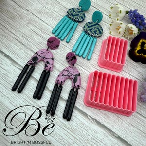 Può includere: Due paia di orecchini fatti a mano in argilla polimerica con design geometrici e astratti. Gli orecchini sono esposti su un espositore per orecchini in plastica rosa. L'espositore ha più fessure per tenere gli orecchini.