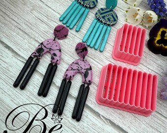 May include: Two pairs of handmade polymer clay earrings with geometric and abstract designs. The earrings are displayed on a pink plastic earring display stand. The stand has multiple slots for holding earrings.