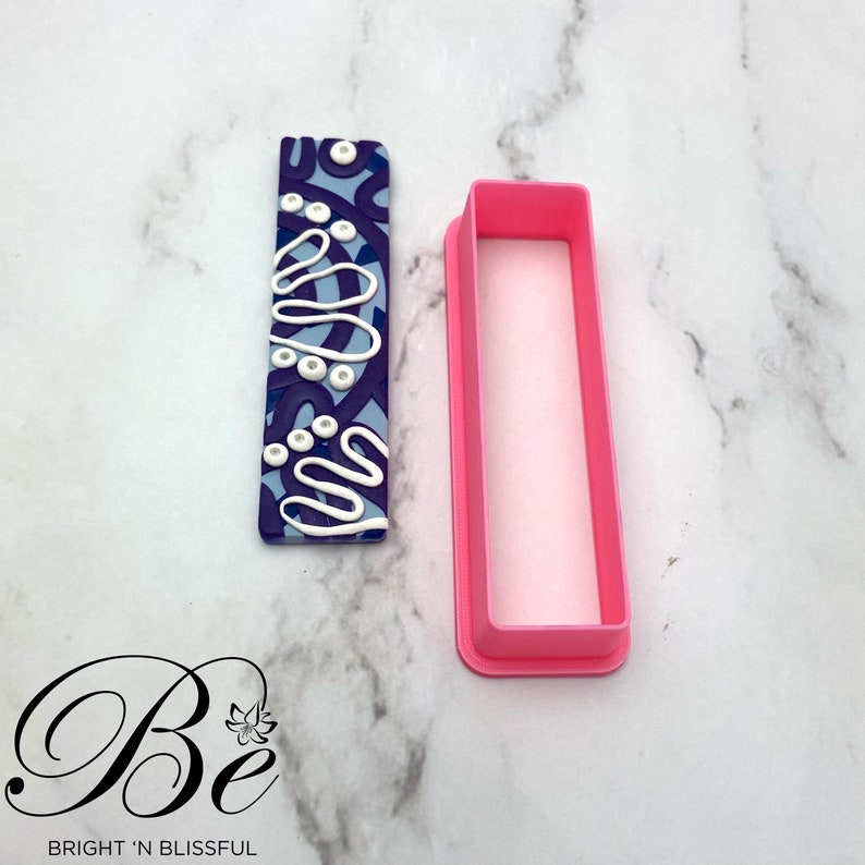 Op de afbeelding: Een roze plastic rechthoekige koekjesuitsteker met een blauw en wit gepatroneerd rechthoekig stukje polymer clay ernaast. Het polymer clay-rechthoekje heeft een wit swirl-ontwerp en witte stippen.