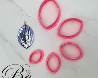 May include: Set of five pink plastic leaf-shaped cookie cutters. The cutters are arranged in a semi-circle on a white surface. A pair of earrings with a purple and silver leaf design is also pictured.