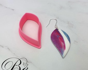 Puede incluir: Un par de pendientes en forma de hoja de color rosa y blanco con vetas marmoleadas y ganchos plateados. Los pendientes se muestran junto a un molde de plástico rosa en forma de hoja.