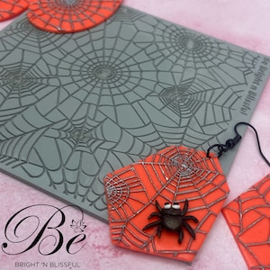 May include: A gray silicone mold with a spiderweb design. The mold is used to create polymer clay earrings. The earrings are orange with a silver spiderweb design and a black spider.