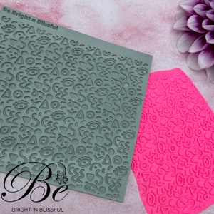 May include: Two silicone baking mats with a repeating pattern of eyes, triangles, and wavy lines. The mats are green and pink. The text "Be Bright 'n Blissful" is printed on the green mat.