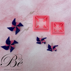 Pode incluir: Brincos de cata-vento rosa e azul com ganchos prateados. Os brincos são exibidos em uma superfície rosa com duas formas de plástico rosa para fazer os brincos.