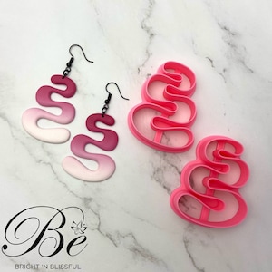 Pode incluir: Um par de brincos rosa degradê com um design ondulado e ganchos pretos. Ao lado dos brincos, há dois cortadores de biscoitos de plástico rosa com o mesmo design ondulado.