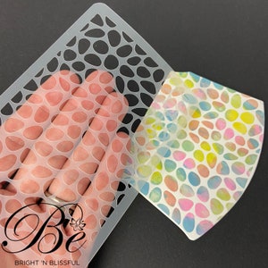 May include: A clear plastic stencil featuring a repeating pattern of small, rounded shapes. The stencil is being positioned over a piece of paper with a similar pattern printed on it in shades of pink, blue, yellow, and green. The paper has the text "Be" and "BRIGHT 'N BLISSFUL" printed on it.