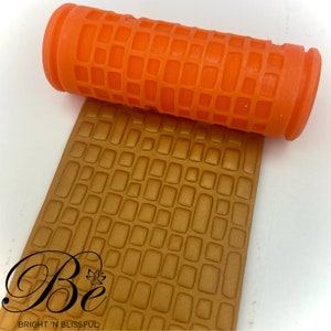 Peut inclure: Rouleau à pâtisserie en silicone orange avec un motif carré. Le rouleau à pâtisserie roule sur une surface brune avec un motif carré en relief. La surface est étiquetée "Be Bright 'N Blissful".