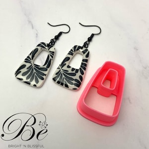 Peut inclure: Une paire de boucles d'oreilles rectangulaires à motif noir et blanc avec une découpe au centre. Les boucles d'oreilles sont suspendues à des crochets noirs. Un moule en plastique rose pour la fabrication des boucles d'oreilles est également présenté.