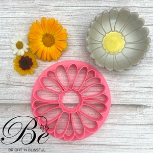 Könnte beinhalten: Rosaer Plastik-Ausstecher in Form einer Daisy mit 12 Blütenblättern. Der Ausstecher liegt auf einer weißen Holzoberfläche mit einem silbernen Daisy-förmigen Ausstecher und drei echten Gänseblümchen.
