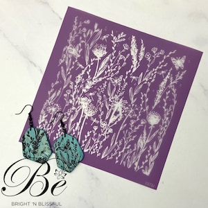 Könnte beinhalten: Ein Paar türkisblaue Ohrringe mit einem floralen Design. Die Ohrringe sind aus Holz und haben einen schwarzen Metallhaken. Die Ohrringe werden auf einem violetten Hintergrund mit einer weißen floralen Schablone gezeigt.