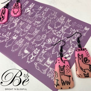 May include: A pair of pink and orange llama earrings with black outlines. The earrings are shaped like a teardrop and have a black hook. The earrings are on a purple background with a white llama pattern.