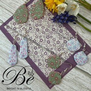 May include: Three pairs of handmade earrings with floral patterns. The earrings are made of polymer clay and have silver hooks. The patterns are in shades of green, blue, pink, and white.