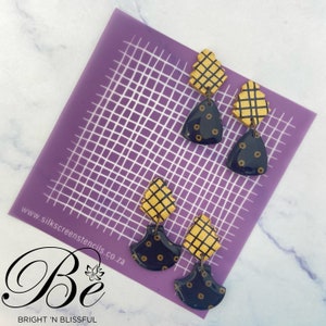 Könnte beinhalten: Ein Paar Ohrringe mit geometrischem Design. Die Ohrringe sind aus schwarzem und goldenem Polymerton gefertigt. Der obere Teil der Ohrringe ist ein Gittermuster und der untere Teil ist eine Fächerform mit goldenen Punkten. Die Ohrringe werden auf einer violetten Gittermatte präsentiert.