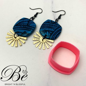 May include: A pair of blue and black patterned earrings with gold sunburst accents. The earrings are displayed on a white surface with a pink square-shaped container.