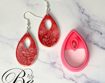Peut inclure: Une paire de boucles d'oreilles en forme de larme rouge et blanche avec un motif découpé. Les boucles d'oreilles sont en résine et ont un crochet argenté. Un moule en silicone rose pour la fabrication des boucles d'oreilles est également présenté.