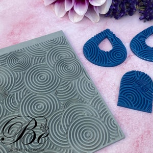 May include: A gray silicone mold with a repeating pattern of concentric circles. Three blue resin pieces are shown, each with the same concentric circle pattern. The resin pieces are shaped like a teardrop, a heart, and a rounded rectangle. The mold is labeled "Be Bright 'n Blissful".
