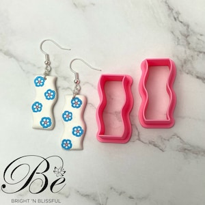 Puede incluir: Pendientes blancos con forma de onda con diseños florales azules y rosas, colgados de ganchos plateados. También hay dos cortadores de arcilla rosas con forma de onda. Los pendientes y cortadores están sobre una superficie de mármol blanco.