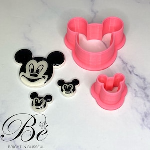 May include: A collection of Mickey Mouse-themed cookie cutters and decorative pieces. The cutters are bright pink, and the decorative pieces feature black and white Mickey Mouse faces. The logo "Bright 'N Blissful" is visible in the bottom left corner.