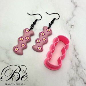 Peut inclure: Une paire de boucles d'oreilles en argile polymère rose avec un motif ondulé et des accents de fleurs blanches. Les boucles d'oreilles sont suspendues à des crochets noirs. Un moule en plastique rose avec un motif ondulé est également présenté.