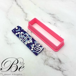 Op de afbeelding: Een roze plastic rechthoekige koekjesuitsteker met een blauwe en witte gepatroneerde polymer clay-staaf erin.