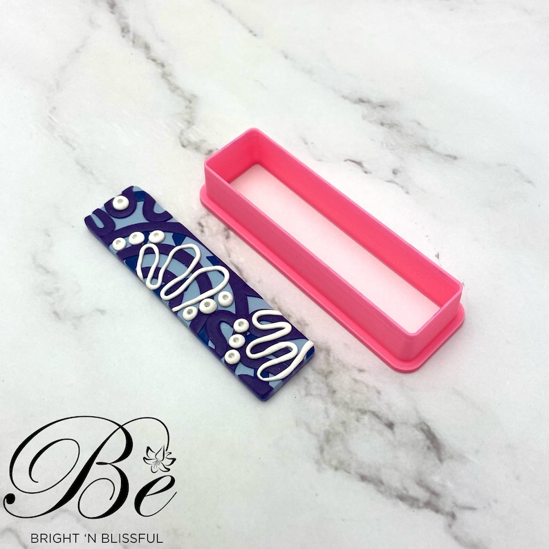 Op de afbeelding: Een roze plastic rechthoekige koekjesuitsteker met een blauwe en witte gepatroneerde polymer clay-staaf erin.