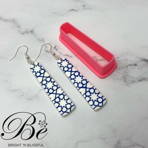 Op de afbeelding: Een paar hangende oorbellen met een witte achtergrond en een blauw geometrisch patroon. De oorbellen zijn zilverkleurig en hebben een haaksluiting. De oorbellen worden gepresenteerd op een roze achtergrond.
