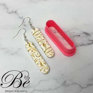 Può includere: Un paio di orecchini pendenti bianchi e dorati con un gancio d'argento. Gli orecchini sono realizzati in pasta polimerica e presentano un motivo floreale. Gli orecchini sono mostrati accanto a uno stampo in plastica rosa.