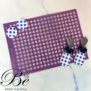 May include: A pair of dangle earrings with a white base and blue floral pattern. The earrings are made of polymer clay and have a silver and black accent. The earrings are displayed on a purple background with a white floral pattern.